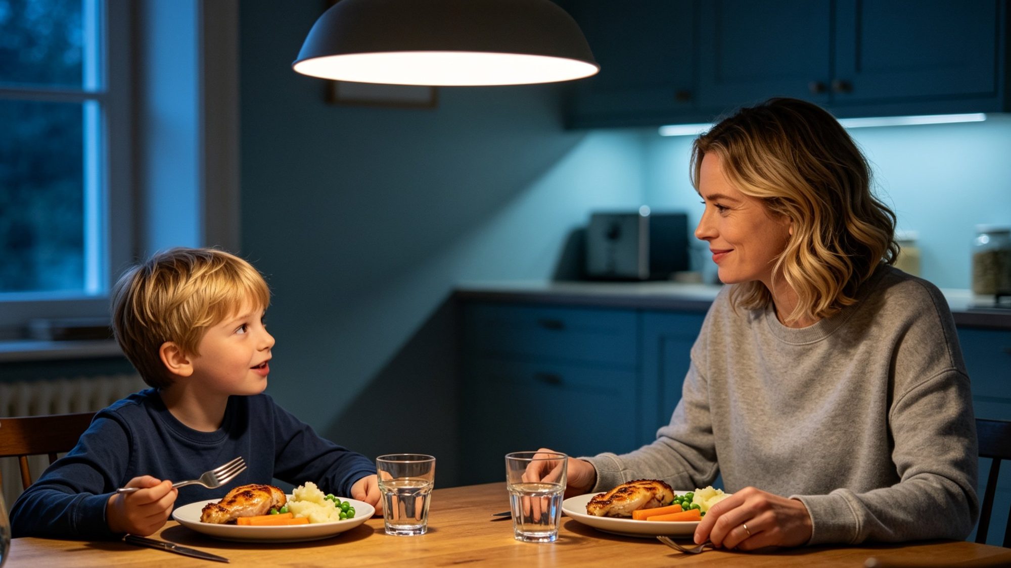 A mother and her young son sharing a weeknight dinner at the dining table at home. Sensible meat-and-three-veg meal, warm pendant light overhead, the boy mid-conversation, the mother listening.