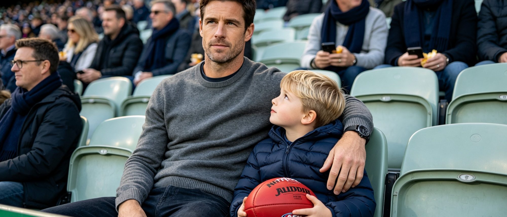 A father at an AFL match at the MCG with his young son leaning into him, the boy looking up at his dad while the father gazes thoughtfully into the middle distance.